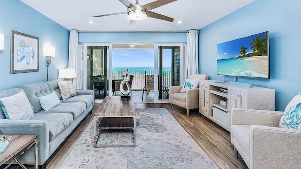 A spacious living room with a large window, a ceiling fan, and a flat-screen TV. - On the Sand! Beachfront Pool & Views! Walk to Everything! (Fort Myers Beach)