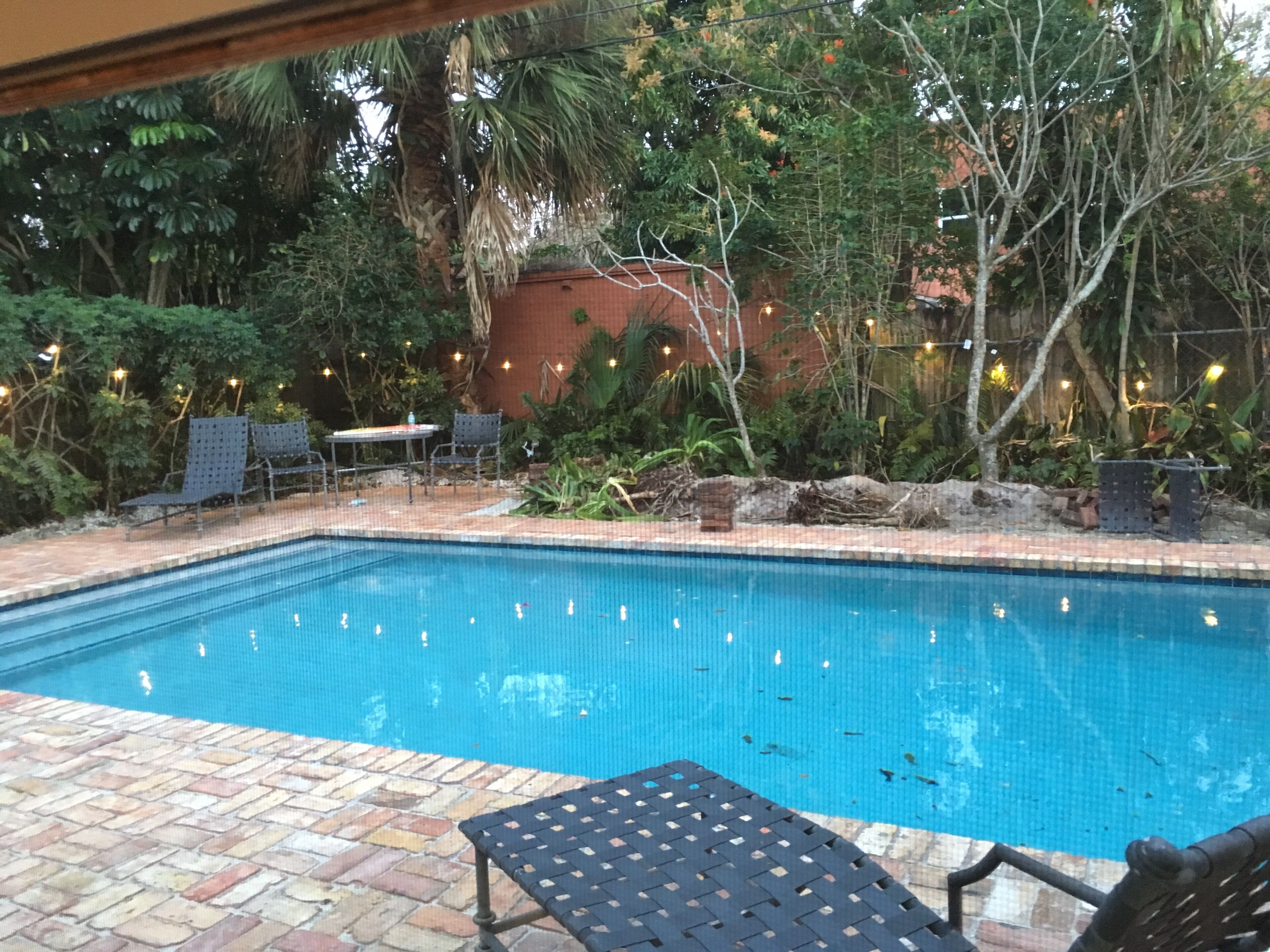 Uma piscina retangular com borda azulejada, rodeada por um terraço de tijolos. Há duas cadeiras e uma pequena mesa perto da piscina.