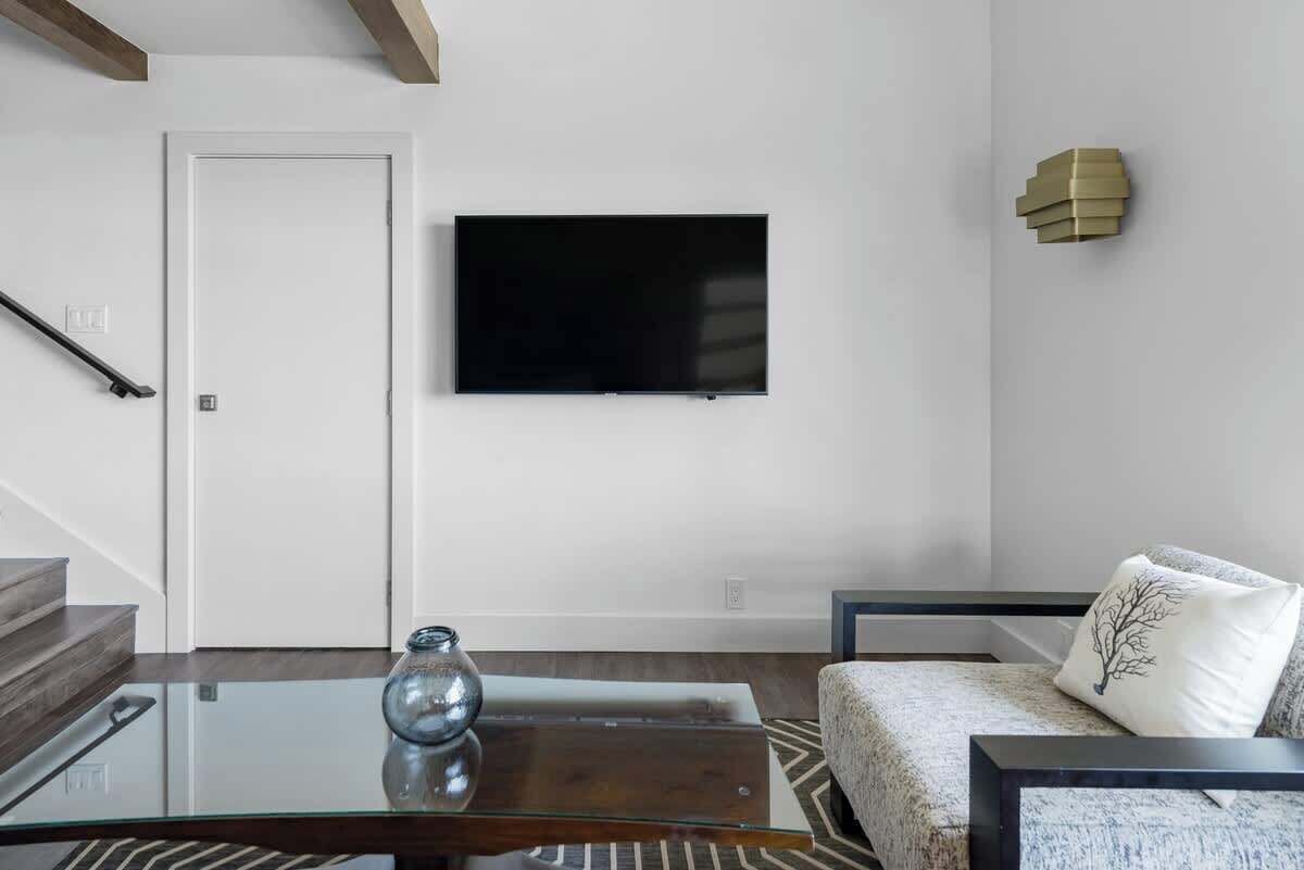 A modern living room with a glass coffee table, a sofa, a flat-screen TV mounted on the wall, and a staircase with wooden beams.