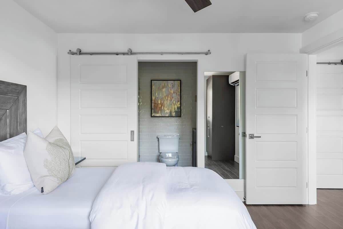 A bedroom with a bed, a white door, and a bathroom visible through an open door.