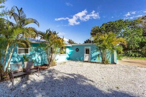 A small, single-story building with a turquoise exterior, a gravel driveway, and several palm trees.