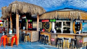 A beachside bar with a thatched roof, stools, and a sign reading "TO GO".