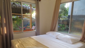 Un lit avec du linge de lit blanc et deux serviettes, une fenêtre donnant sur un toit de chaume et sur la verdure, et des rideaux à la fenêtre.
