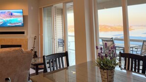 Una sala de estar moderna con televisor, chimenea, comedor con mesa de vidrio y vista al océano a través de amplios ventanales.