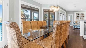 Un coin repas avec une table à plateau de verre, entourée de chaises en osier, de grandes fenêtres et une vue sur l’extérieur.