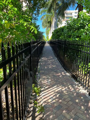 A paved pathway flanked by black metal fences, leading towards a building with a white facade and balconies.