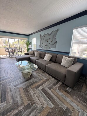A modern living room with a herringbone wood floor, a brown leather sofa, a glass coffee table, and a large wall art piece.