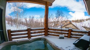 Une terrasse en bois avec un jacuzzi, un sol recouvert de neige et une vue sur un village avec ses bâtiments et ses arbres.