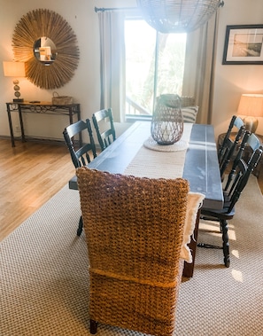 A dining area with a long table, chairs, a woven chair, a wall-mounted mirror, and a window with curtains.