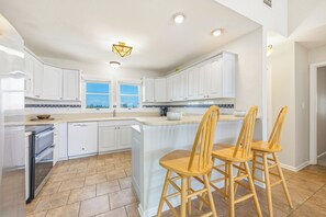 A kitchen with white cabinets, a central island with bar stools, and a view through the windows. - Semi-Oceanfront Home w/ Private Pool, Hot Tub, Game Room, Community Tennis (Waves)