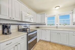 A kitchen with white cabinets, a stove, an oven, a coffee maker, and a window with a view of houses.
