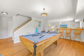 Une table de billard dans une pièce dotée d’un plancher en bois, d’un escalier et d’un coin salon avec des chaises en bois.