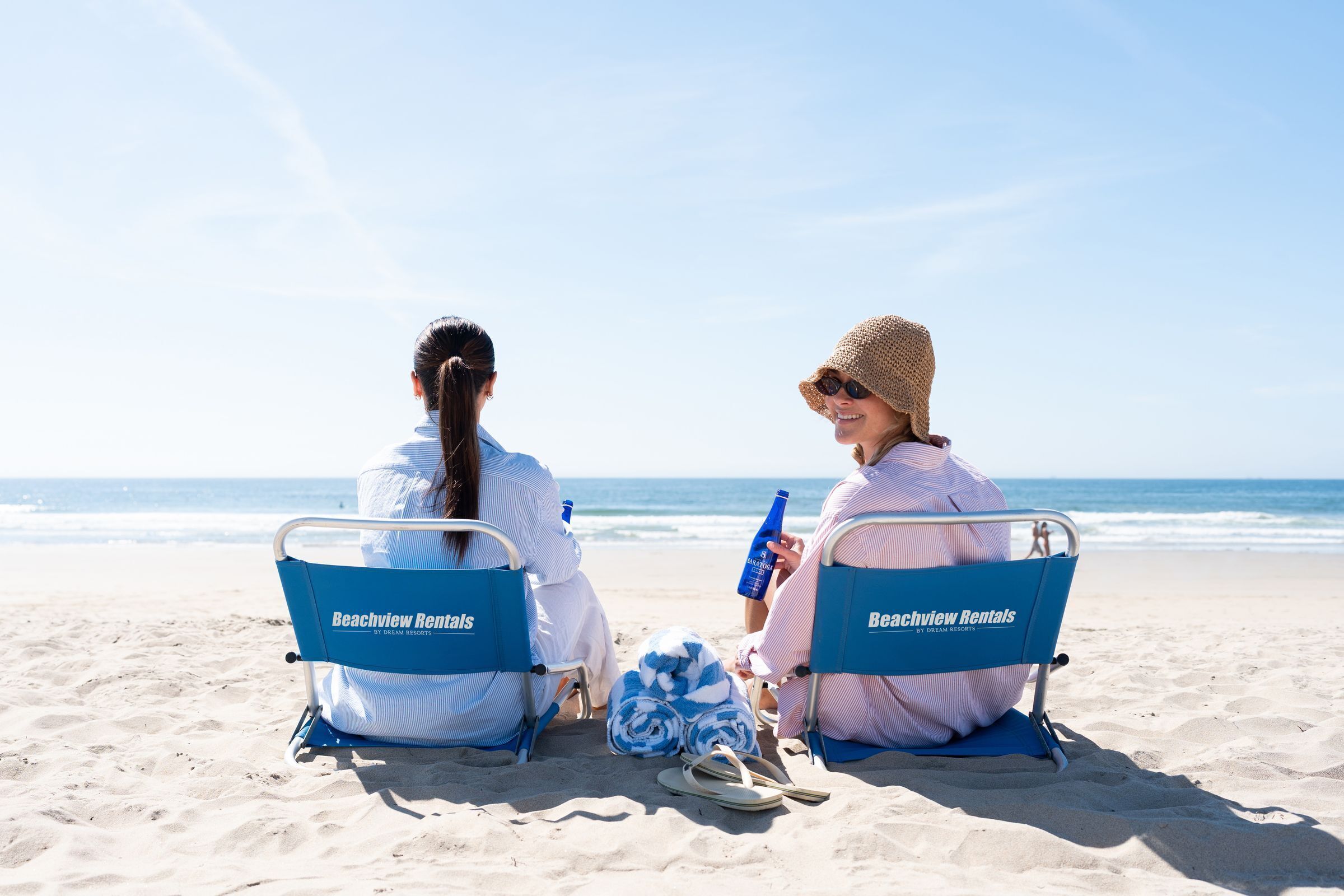 Dos personas sentadas en sillas de playa con la marca 'Beachview Rentals', en una playa arenosa con el océano al fondo.
