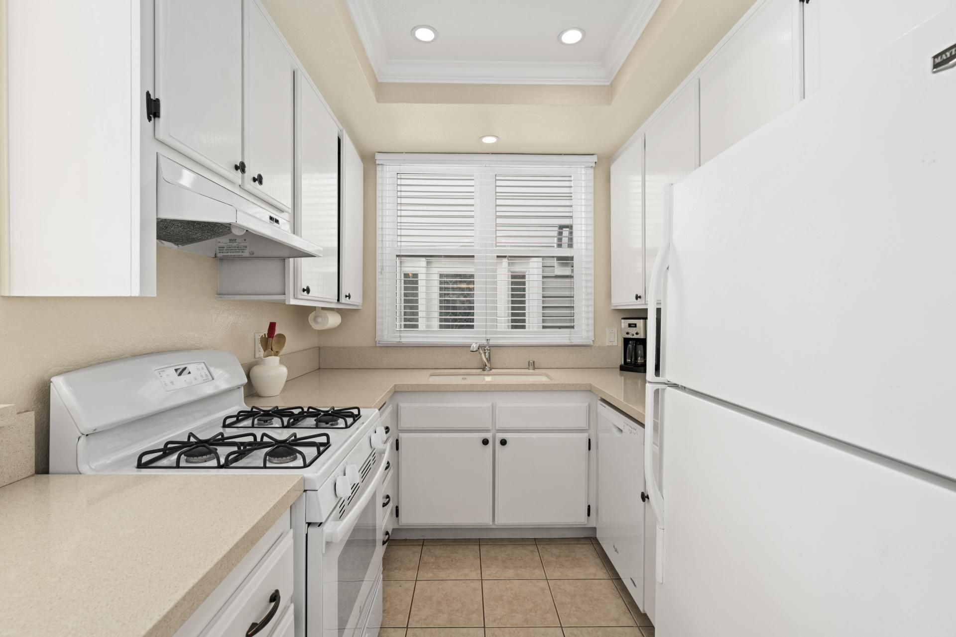 A compact kitchen with white cabinets, a gas stove, a refrigerator, and a window with blinds.