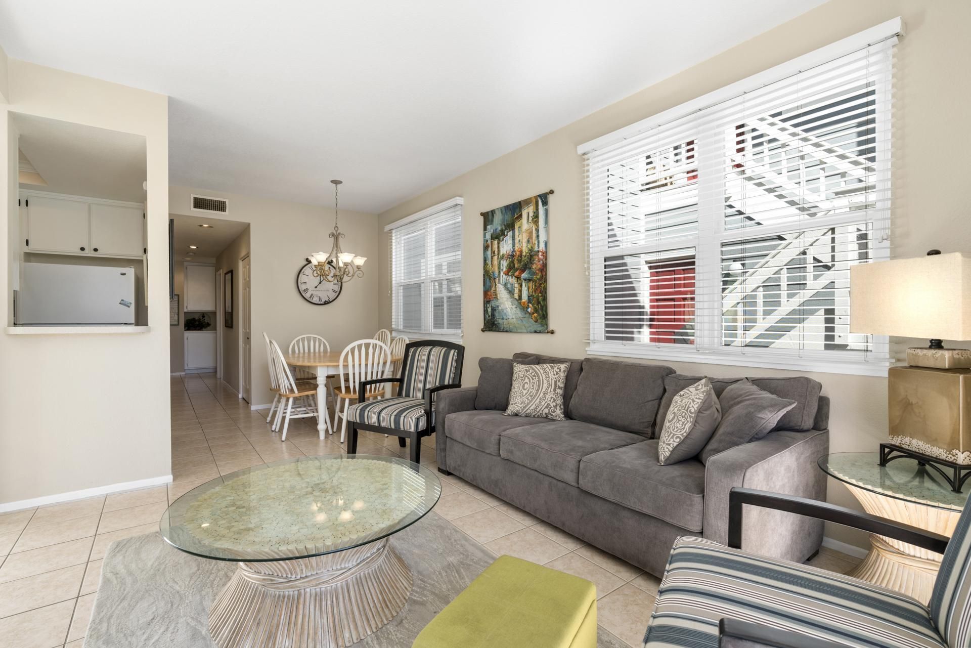 A living room with a gray sofa, a glass coffee table, and striped chairs. There is a painting on the wall and a clock above the fireplace.