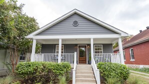 Une maison à deux étages avec un porche à l’avant, une porte d’entrée et un petit jardin devant, agrémenté d’arbustes et d’un chemin menant à la porte.