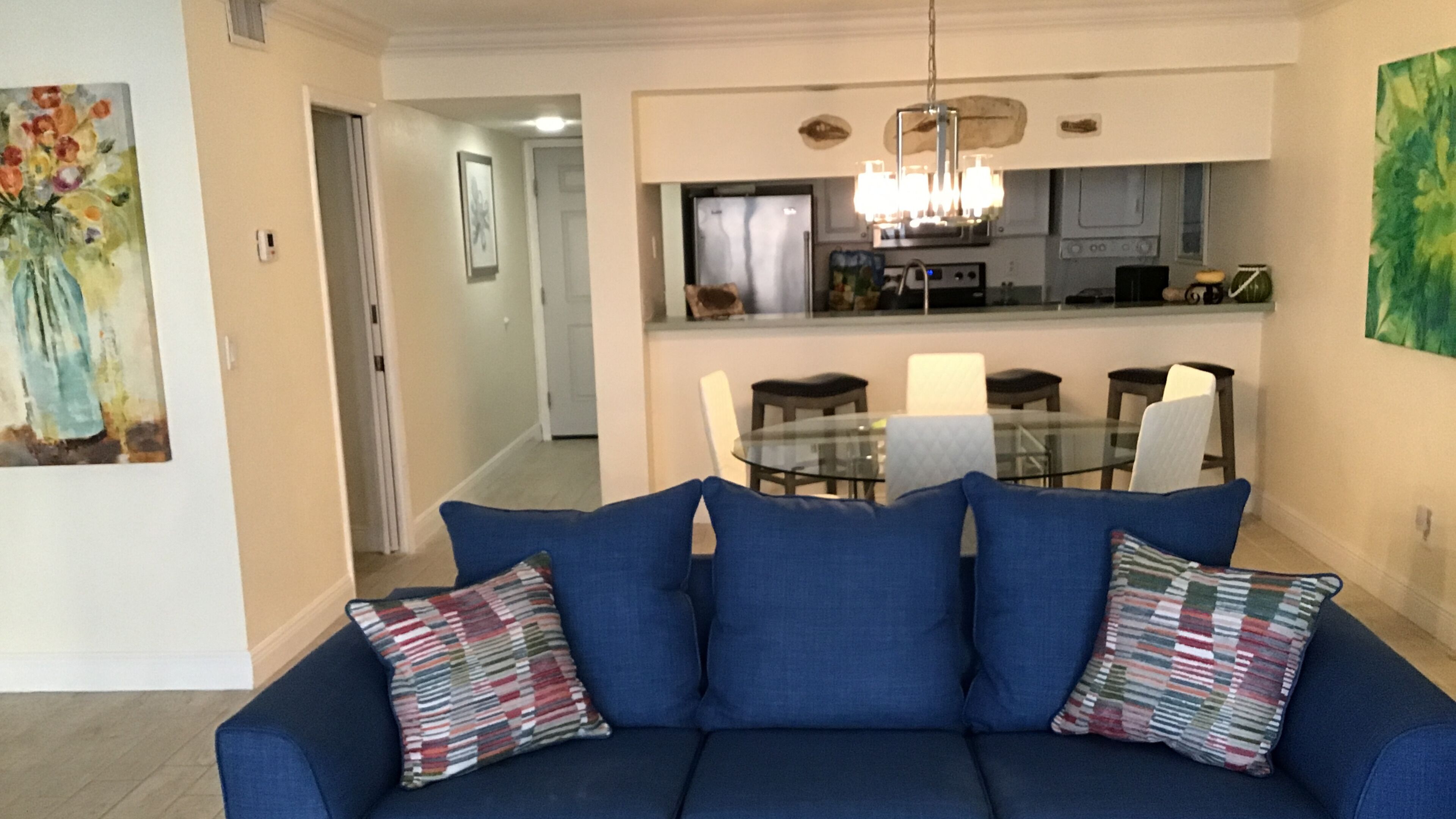 Uma sala de estar com um sofá azul, uma mesa de jantar de vidro e uma cozinha ao fundo.