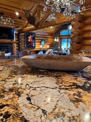 Un intérieur rustique avec un plafond en bois, un grand saladier en bois posé sur un plan de travail en granit et une tête de cerf accrochée au mur.