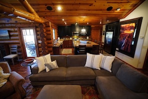L’intérieur d’une cabane en rondins présente un salon avec un grand canapé d’angle, un coin repas avec une cuisine en arrière-plan et une cheminée.
