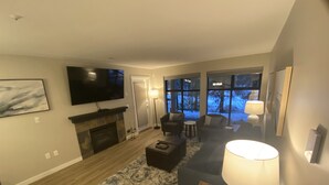 A modern living room with a fireplace, comfortable seating, a flat-screen TV, and large windows offering a view of a snowy landscape.