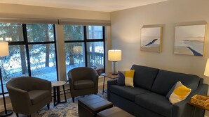 A modern living room with a grey sofa, two armchairs, a coffee table, and a side table with a lamp. Large windows offer a view of trees and a deck outside.