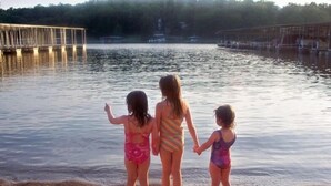 Trois enfants se tiennent debout sur une plage de sable, se tenant la main, face à une étendue d’eau calme, avec une jetée en bois en arrière-plan.