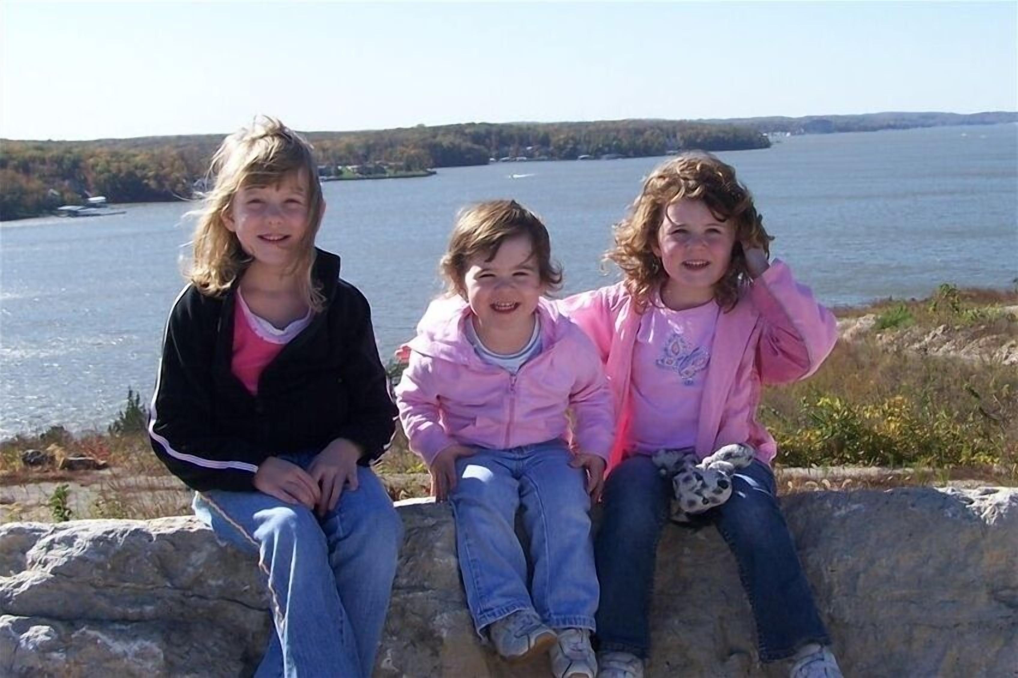 Three children sitting on a stone ledge by a lake.