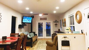 A well-lit living room with a TV, a sofa, a dining table with chairs, a ceiling fan, and a door labeled "PARADISE".