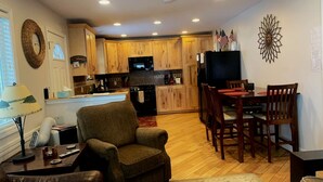 A cozy living room with a sofa, a coffee table, and a TV. There is a dining area with a table and chairs. The kitchen has wooden cabinets and a refrigerator.
