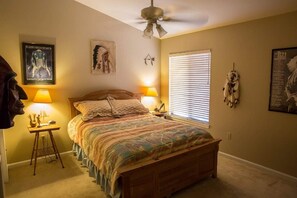 A bedroom with a bed, two bedside lamps, a ceiling fan, a window with blinds, and wall decorations including a poster and a dreamcatcher.