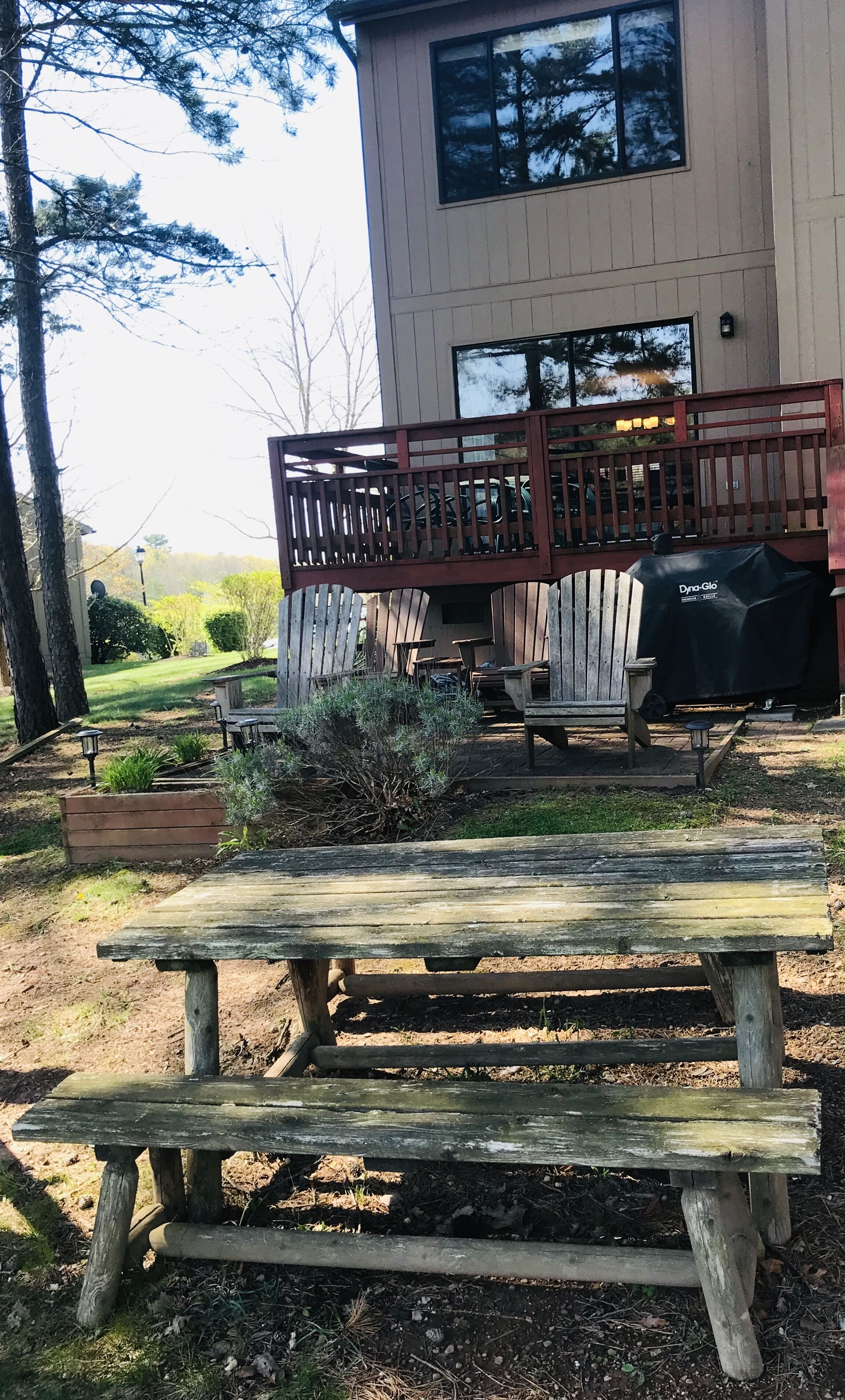 Una terraza de madera con una parrilla, mobiliario exterior y un edificio con grandes ventanales al fondo.