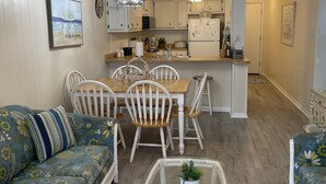 Una sala de estar acogedora con sofá, mesa de comedor y sillas, una cocina con armarios blancos y un refrigerador, y un ventilador de techo.