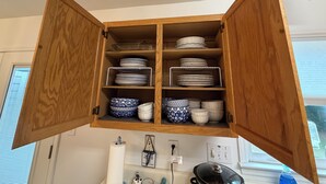 Un buffet en bois avec des étagères où sont rangées des assiettes et des bols, une fenêtre avec des stores et un plan de travail de cuisine avec une bouilloire.