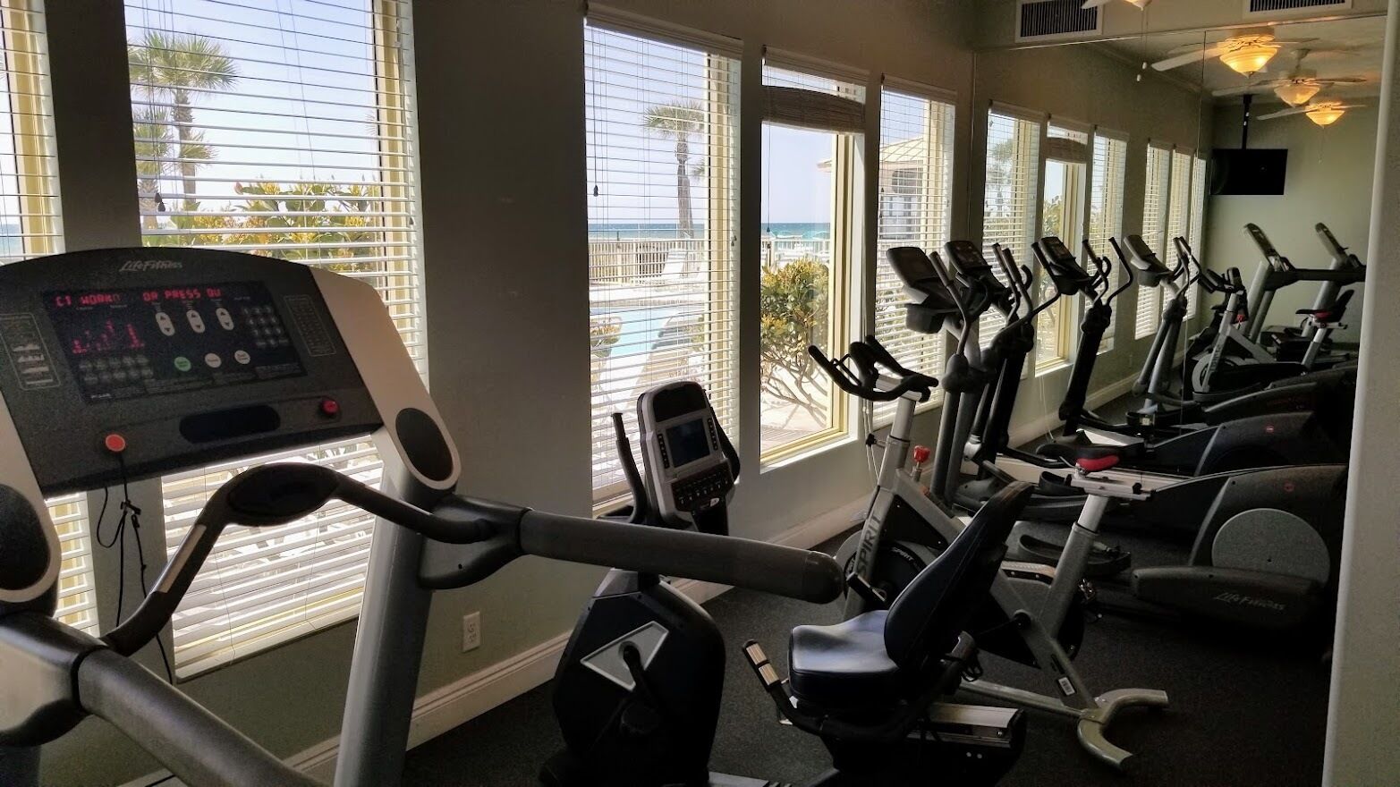 A gym with multiple exercise bikes and a cardio machine, large windows with blinds, and a view of the ocean.