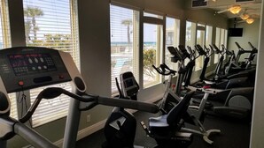 A gym with multiple exercise bikes and a cardio machine, large windows with blinds, and a view of the ocean.