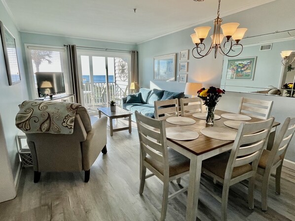 A dining area with a wooden table set for four, surrounded by white chairs. A chandelier hangs above the table, and a sofa with a blue cushion is visible in the background. - APRIL & MAY DISCOUNTED WKS !!NEWLY RENOVATED!! DIRECT OCEAN FRONT CONDO-Unit 104 (Myrtle Beach)