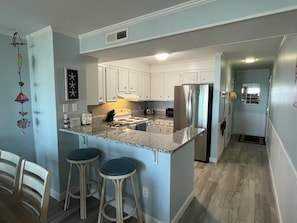 A kitchen with white cabinets, a stainless steel refrigerator, and a granite countertop. There are two bar stools and a wooden chair. -  !!NEWLY RENOVATED!! DIRECT OCEAN FRONT CONDO-Unit 104 (Myrtle Beach)