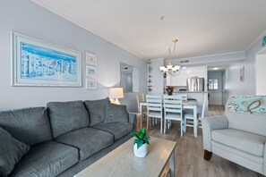 A modern living room with a grey sofa, a coffee table with a plant, and a dining area with white chairs and a table. - APRIL & MAY DISCOUNTED WKS !!NEWLY RENOVATED!! DIRECT OCEAN FRONT CONDO-Unit 104 (Myrtle Beach)