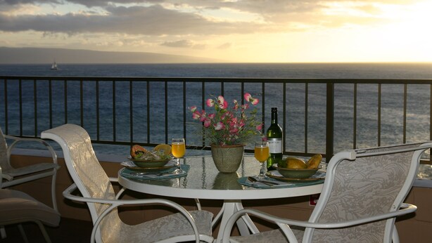 Een ronde tafel met een bloempot, twee glazen sinaasappelsap, een fles wijn en een fruitschaal op een balkon met uitzicht op zee.