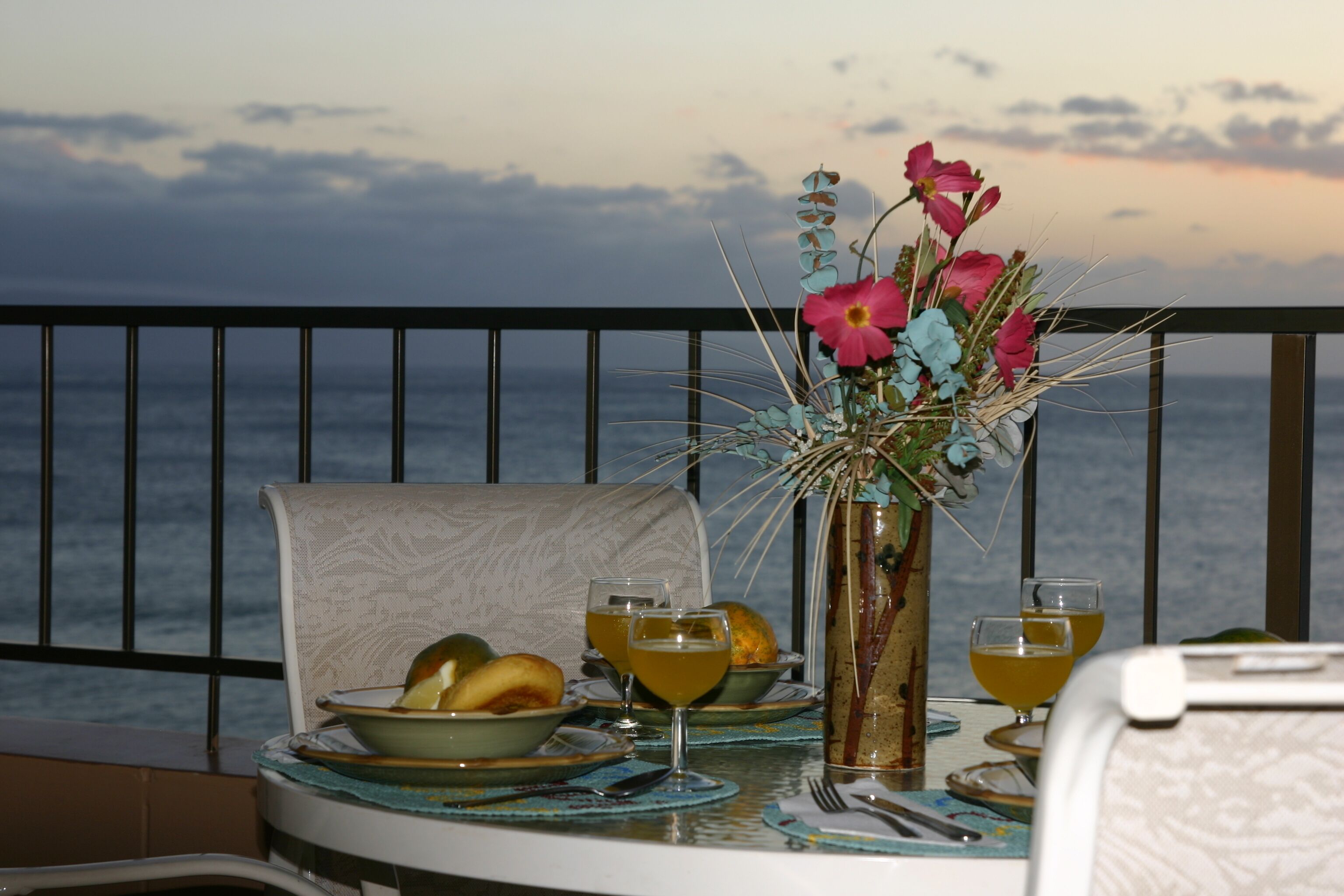 Uma mesa posta com pratos, tigelas e copos de uma bebida amarela, um vaso com flores, e uma cadeira em uma varanda com vista para o mar.