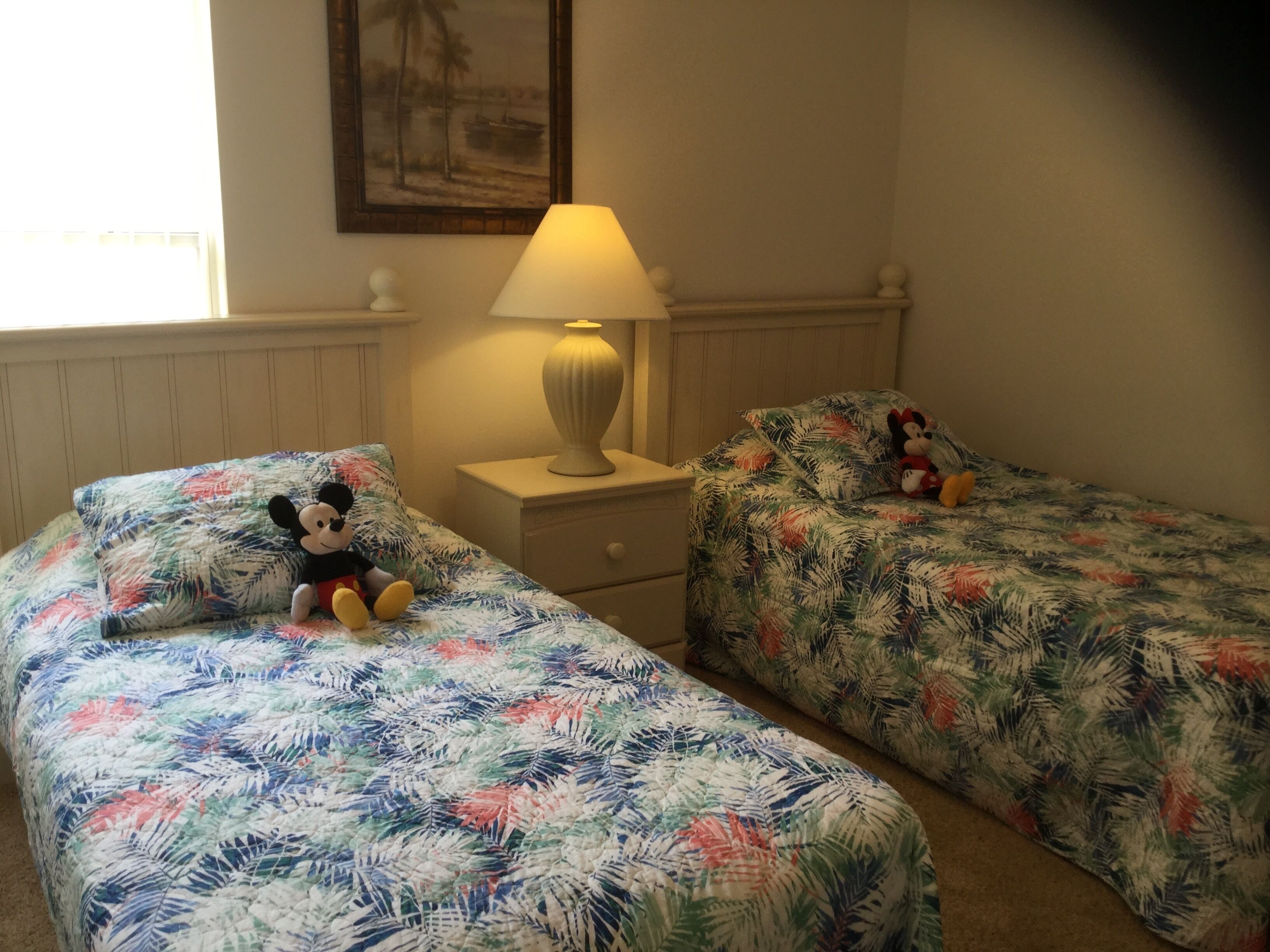 A room with two beds, each with a floral bedspread, a nightstand with a lamp, and a Mickey Mouse plush toy on one bed.