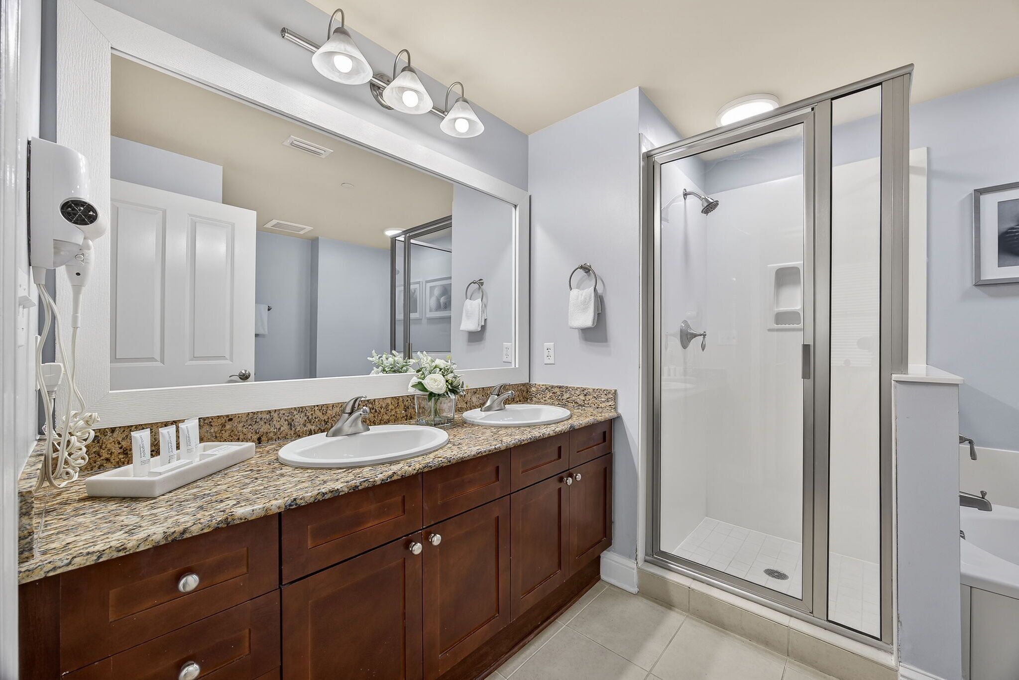 A bathroom with a double sink vanity, a large mirror, a shower enclosure, and a hairdryer mounted on the wall.