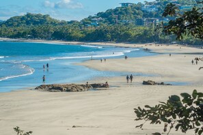 A beach with people walking and standing, a clear sky, and buildings in the background.