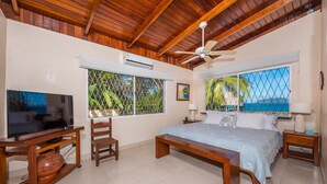A bedroom with a wooden ceiling, a bed with a blue bedspread, a television on a wooden stand, and a window with a view of the sea. - Tamarindo's Hidden Gem, Tropical oasis 3 BR Beachfront Home, with private pool.
 (Tamarindo)
