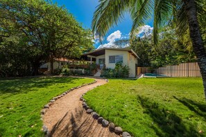 A house with a pathway leading to a pool, surrounded by lush greenery and palm trees.