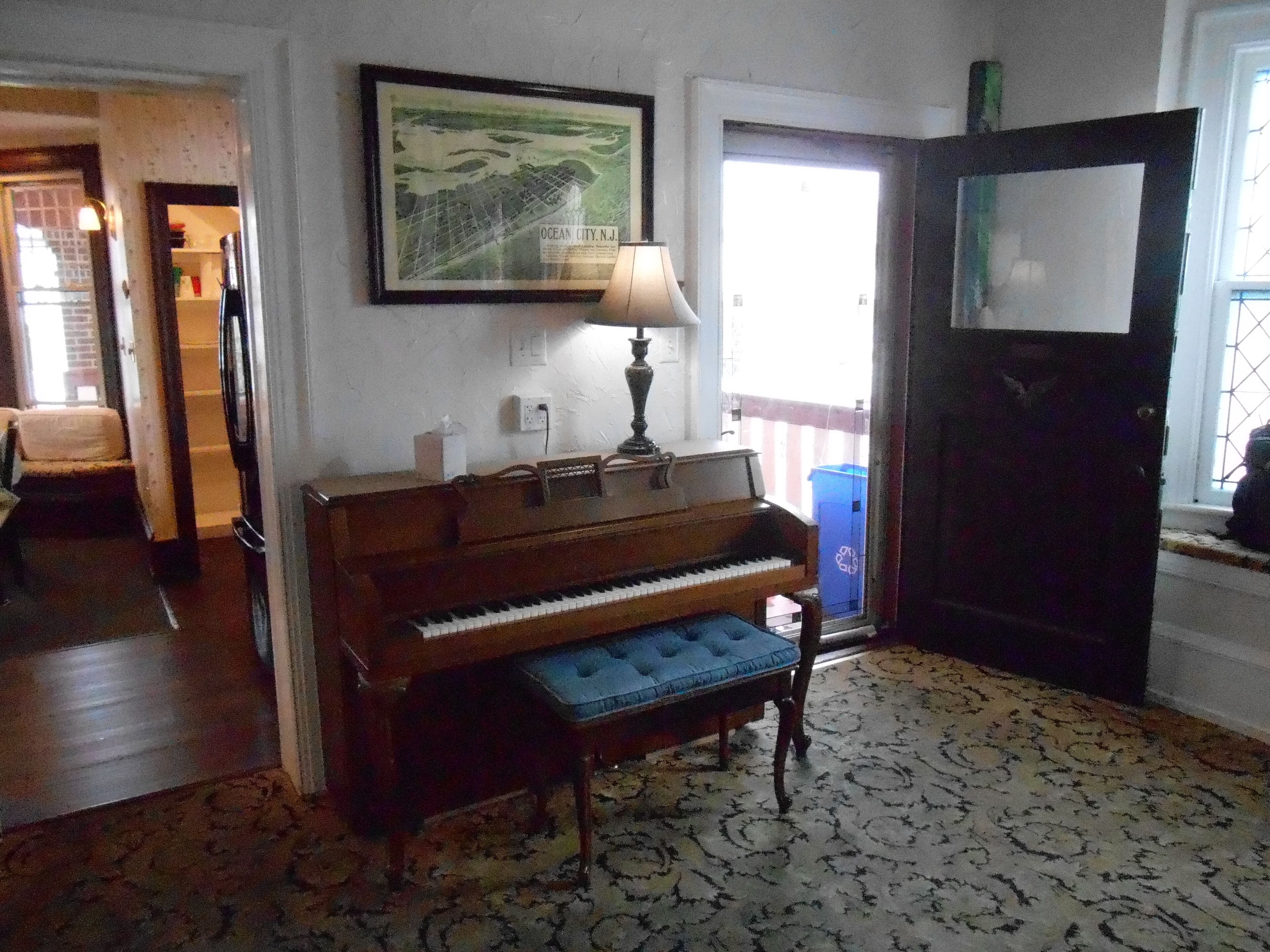 Un piano dans une pièce dont le sol est recouvert d’un tapis à motifs, une carte encadrée accrochée au mur et une porte donnant sur l’extérieur.