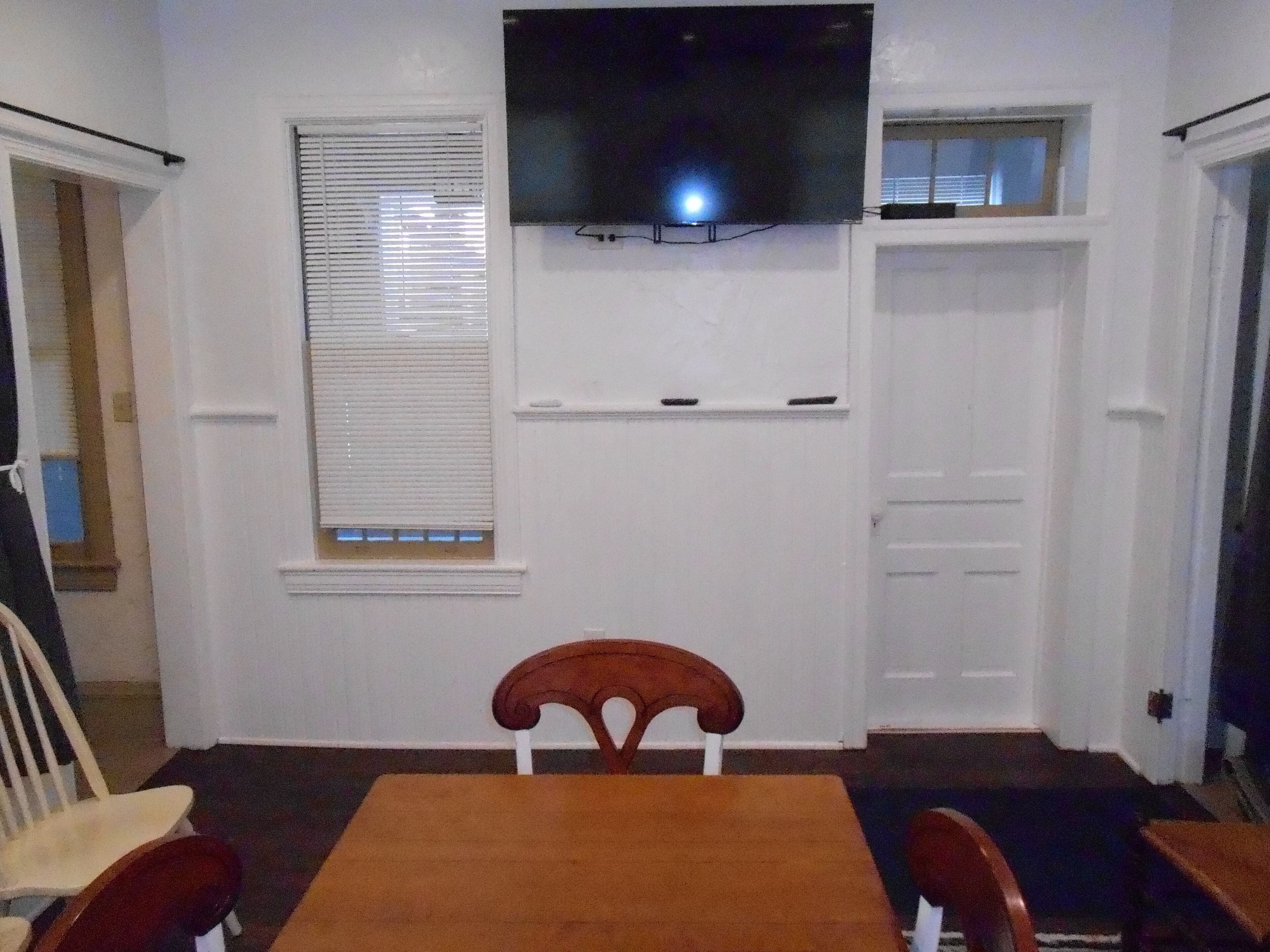Quarto com televisão na parede, janela com persianas, porta branca e mesa de madeira com cadeiras.