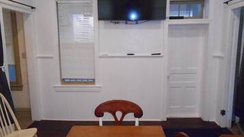 Quarto com televisão na parede, janela com persianas, porta branca e mesa de madeira com cadeiras.