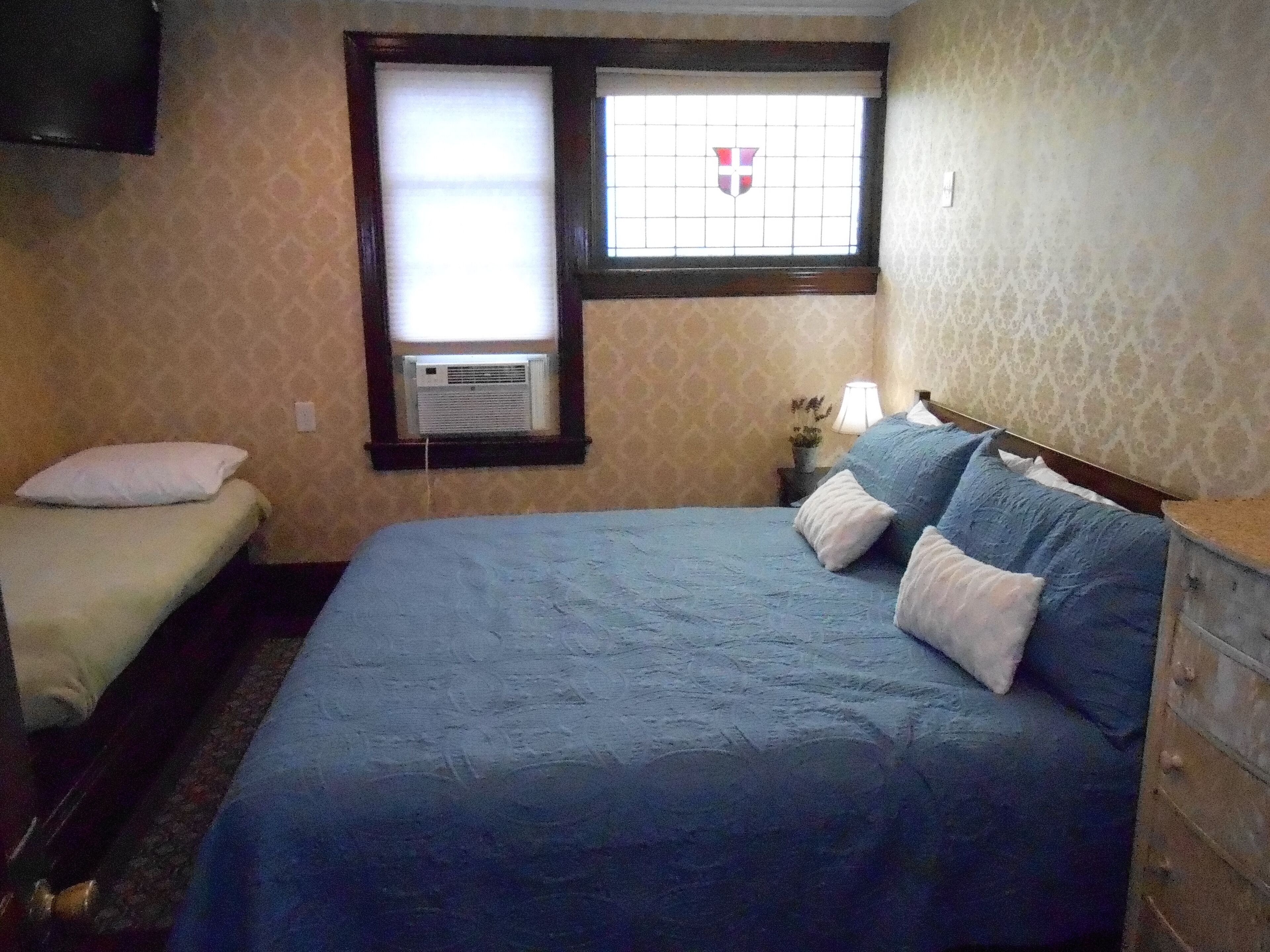 Una camera da letto con un letto grande, un letto più piccolo, una finestra con uno stemma araldico, un comodino con una lampada e un comò.