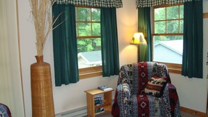 Una sala de estar acogedora con dos ventanas, una lámpara, un sillón, una mesita y un jarrón con plantas secas.